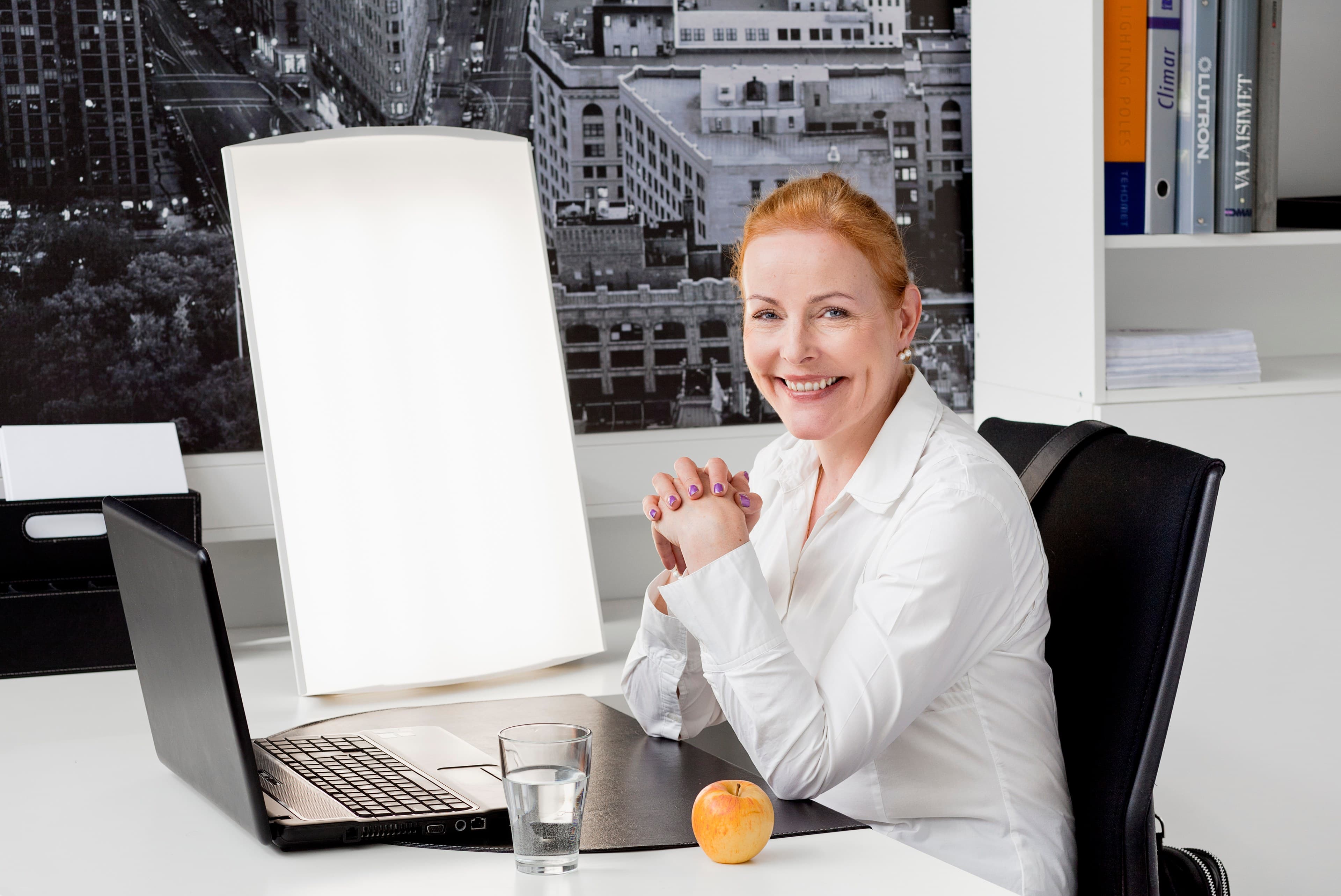 Frau mit roten Haaren sitzt lächelnd am Schreibtisch, links neben ihr leuchtet die Tageslichtleuchte Supernova LED