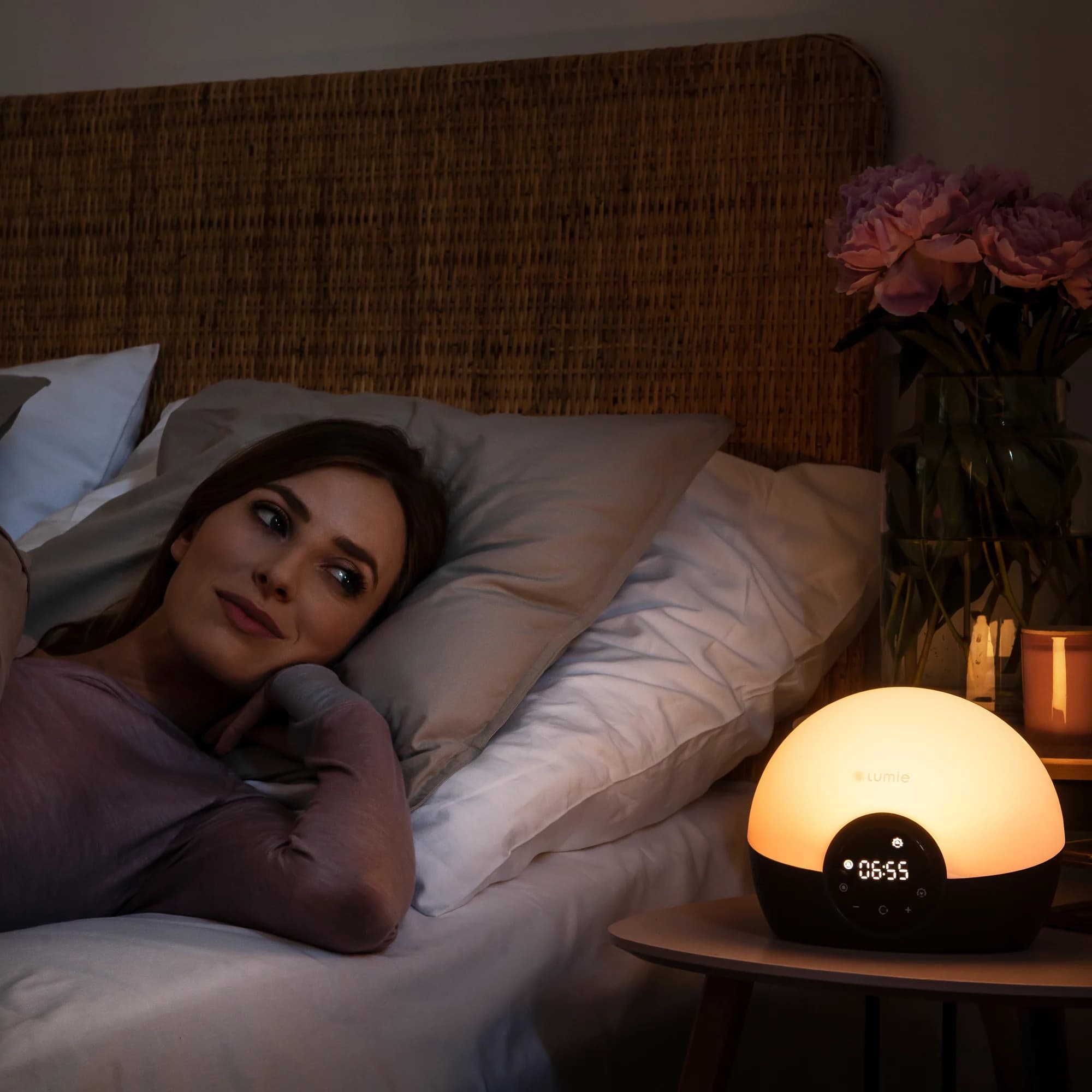 Frau wacht im Bett auf, der Lumie Glow 150 auf dem Nachttisch leuchtet warm, Anzeige 06:55