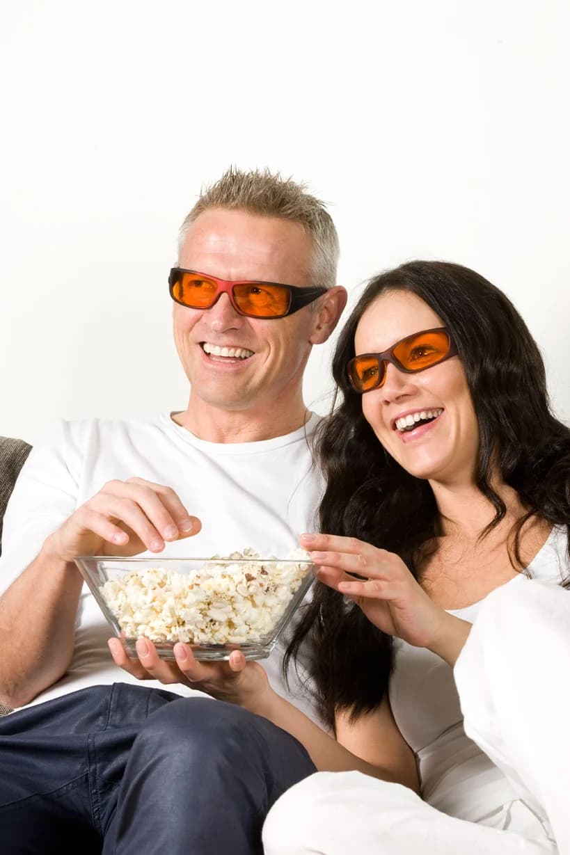 Lachendes Paar sitzt nebeneinander, beide tragen orangefarbene Blueblock-Brillen; der Mann hält eine Glasschale mit Popcorn, die Frau greift hinein.