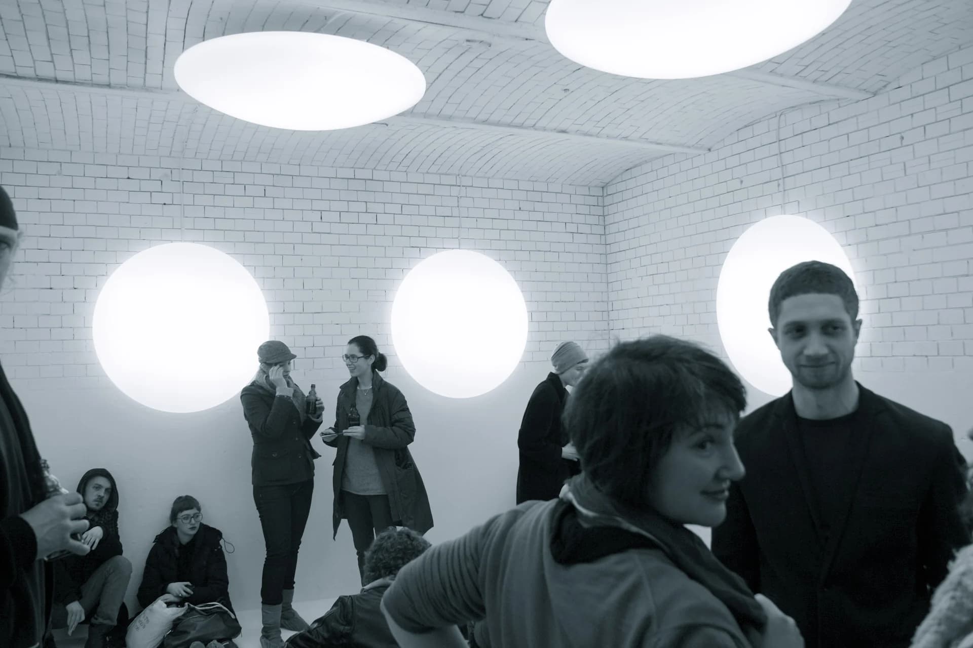 Besucherinnen und Besucher im weiß gestrichenen Gewölberaum der Lichtinstallation in Berlin; an Wänden und Decke leuchten große runde Lichttherapieleuchten.