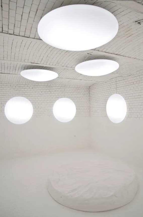 Der leere Lichtraum: weiß gestrichene Ziegelwände und Gewölbedecke mit runden, hell leuchtenden Lichttherapieleuchten, am Boden ein großes rundes weißes Sitzkissen.