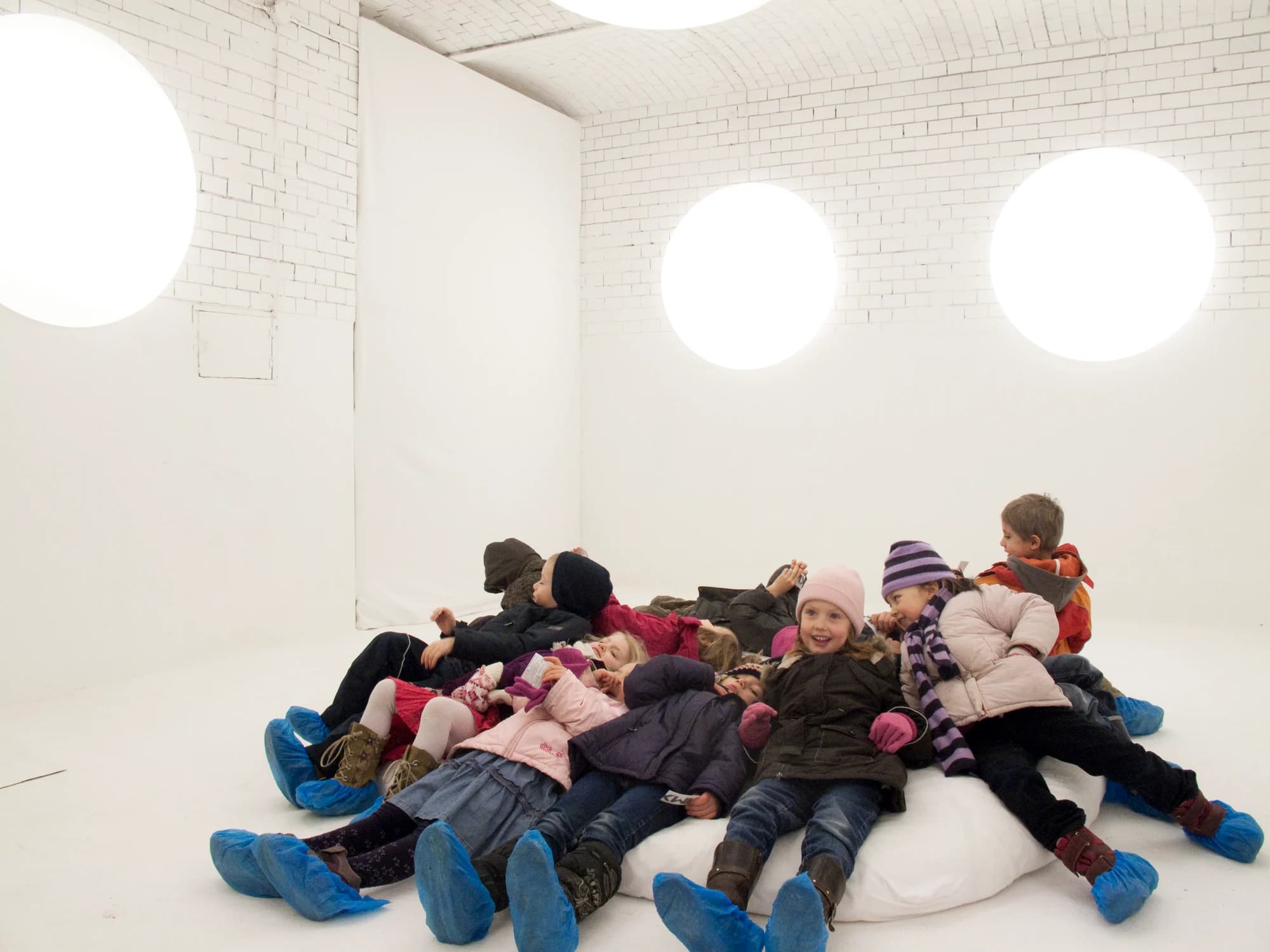 Eine Gruppe Kinder in Winterkleidung und blauen Überziehschuhen liegt und sitzt auf einem großen runden Kissen im weißen Lichtraum; an den Wänden leuchten runde Lichttherapieleuchten.